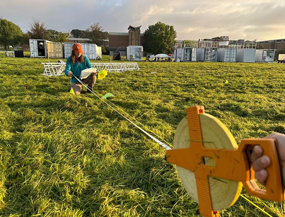 Kneeling on grass, Krista Matuska holds an end of a tape measure while another person, mostly out of the frame, holds the other side.